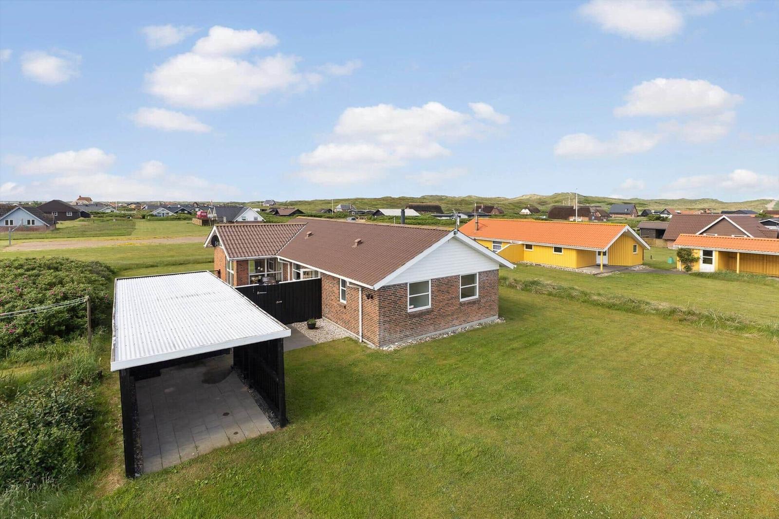 Ein Haus mit Carport auf grünem Grundstück, umgeben von weiteren Häusern und Dünen.