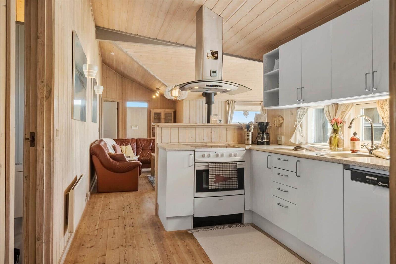 Küche mit weißen Schränken und Holzboden. Im Hintergrund Wohnbereich mit Ledersofa.