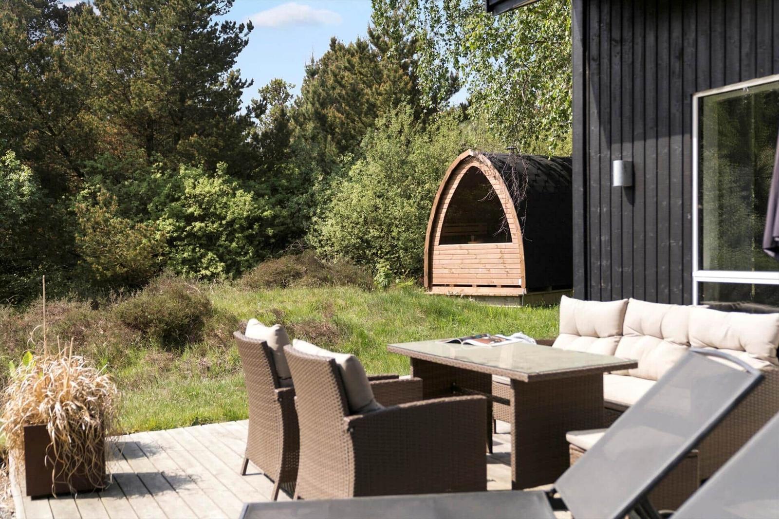 Terrasse mit Sitzgruppe, Tisch und Liegestühlen. Hintergrund: Wald und Holzsauna.