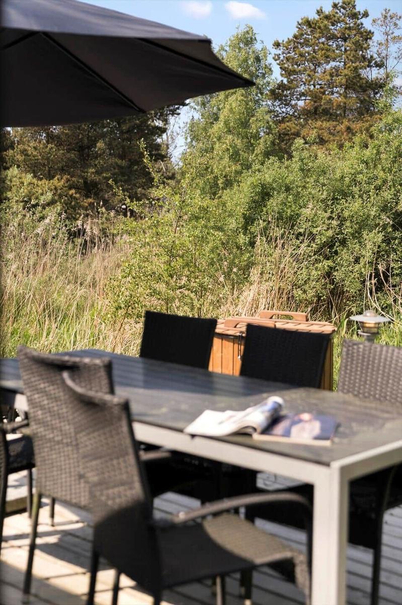 Terrasse mit Tisch, Stühlen und Sonnenschirm. Hintergrund: Grünflächen und Bäume.