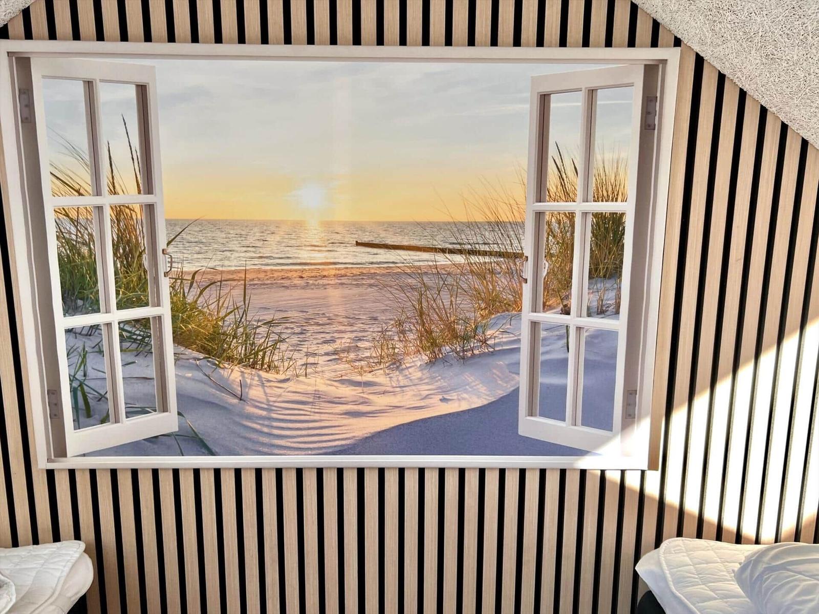Durch das Fenster blickt man auf einen Strand bei Sonnenuntergang mit Dünen und Meer.