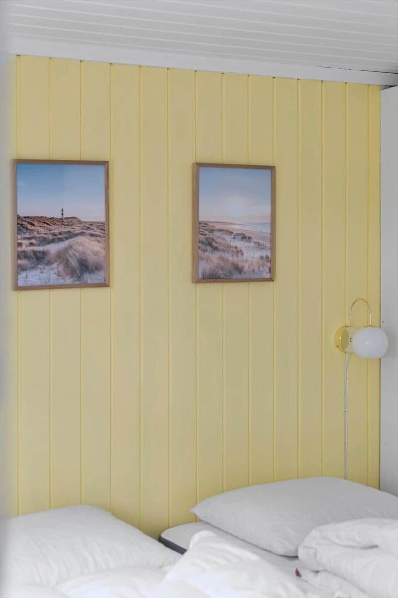 Zwei Betten mit weißen Kissen an einer gelben Paneelwand mit zwei Landschaftsbildern und einer Wandlampe.