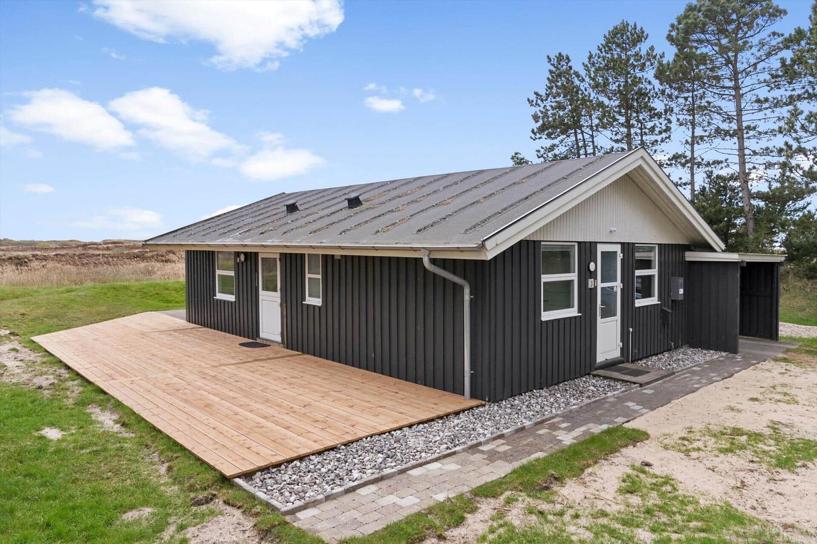 Moderne Hütte mit Holzterrasse und Garten. Weiße Türen und Fenster. Hintergrund mit Bäumen und Gras.