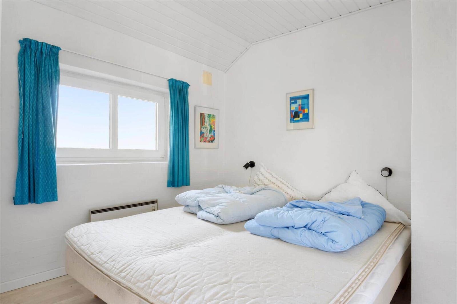 Schlafzimmer mit Bett, blauen Vorhängen und Fenster. An den Wänden hängen Bilder.