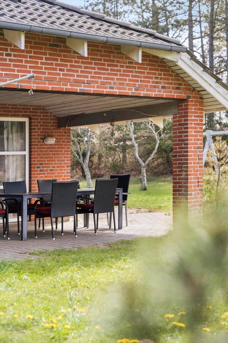 Terrasse mit Tisch und Stühlen unter Dach. Hintergrund: Gras und Bäume.