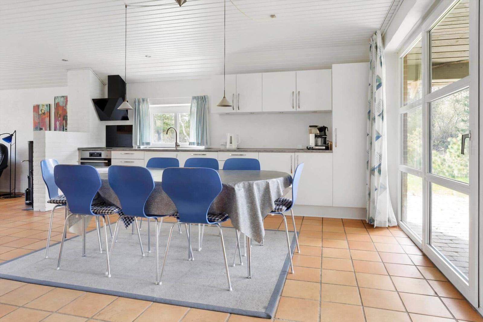 Essbereich mit runder Tafel und blauen Stühlen. Küche mit weißen Schränken. Terrasse über Glastür.