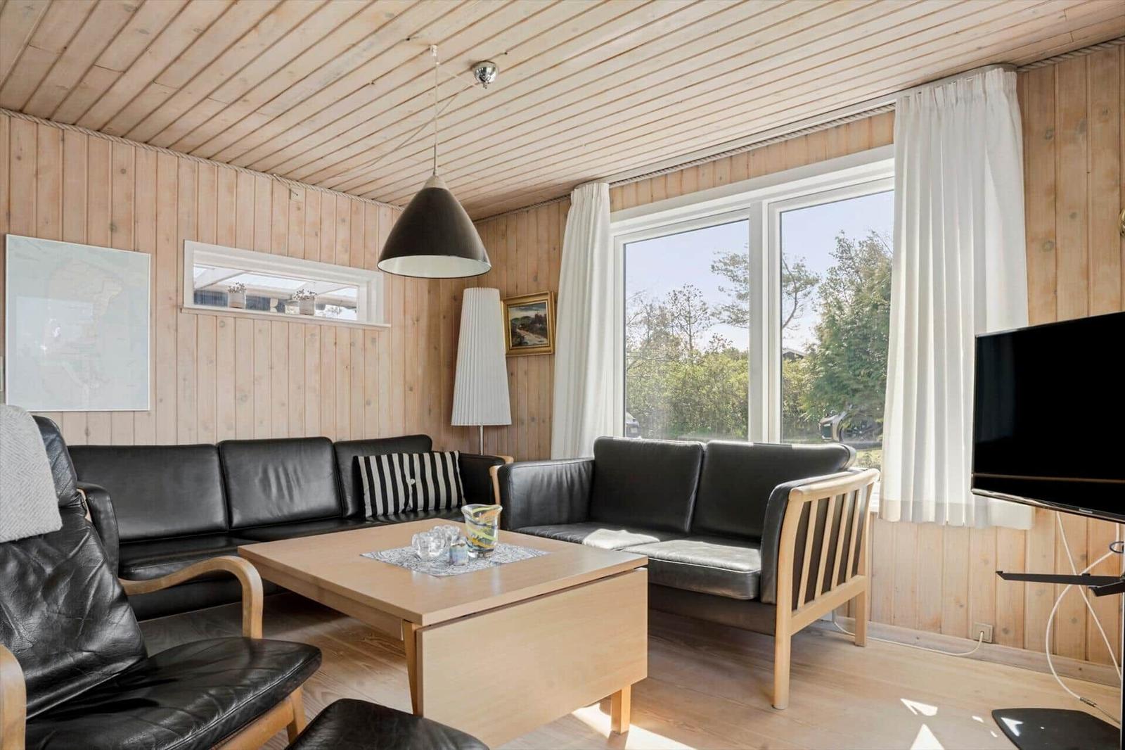Wohnzimmer mit Holzwand, Sofa, Couch und Fernseher. Große Fenster mit Blick nach draußen.