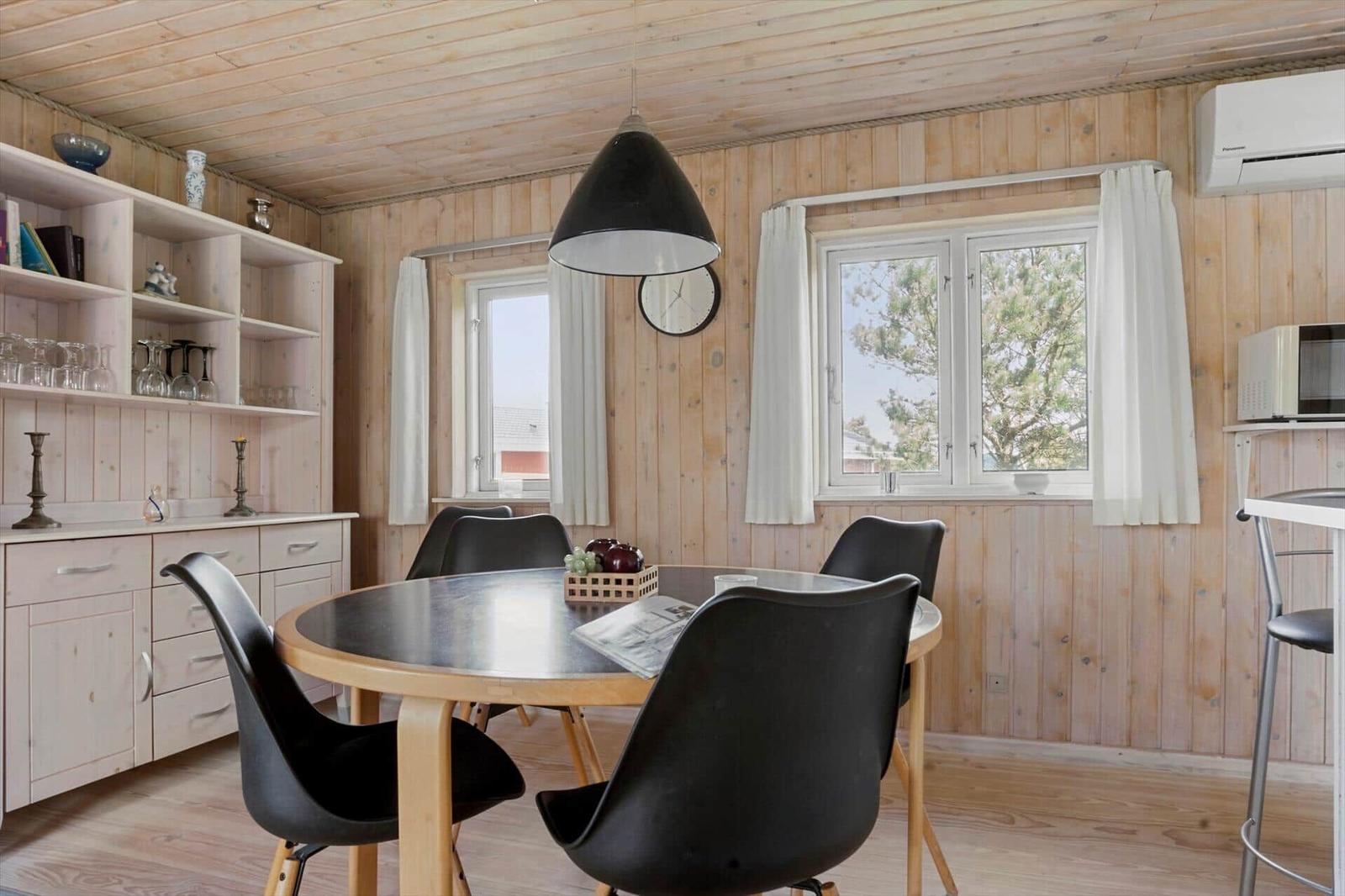 Esszimmer mit runden Tisch, schwarzen Stühlen und Holzwänden. Fenster mit weißen Vorhängen.