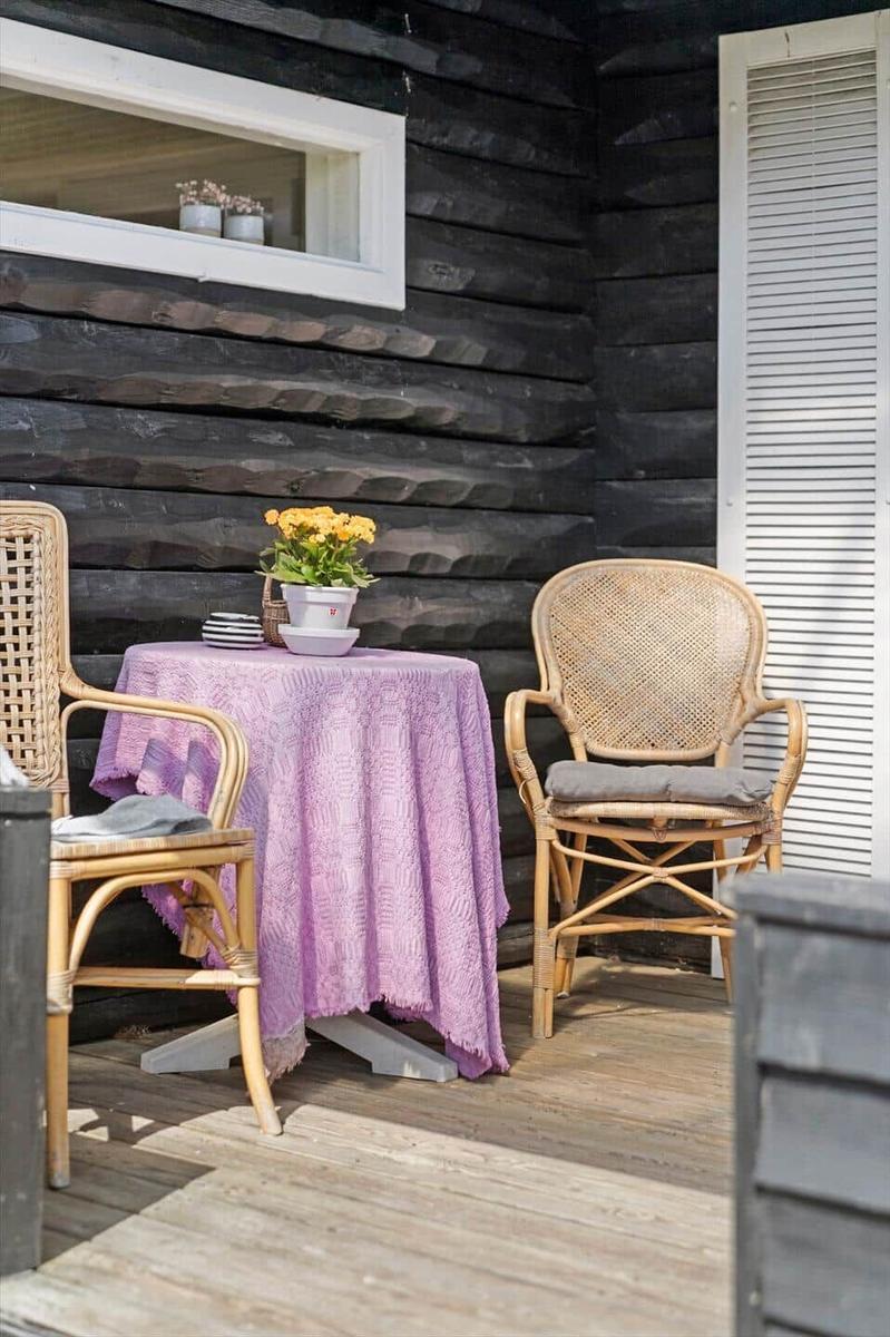 Terrasse mit Tisch, zwei Stühlen und Blumen. Schwarze Holzwand und Holzdielen.