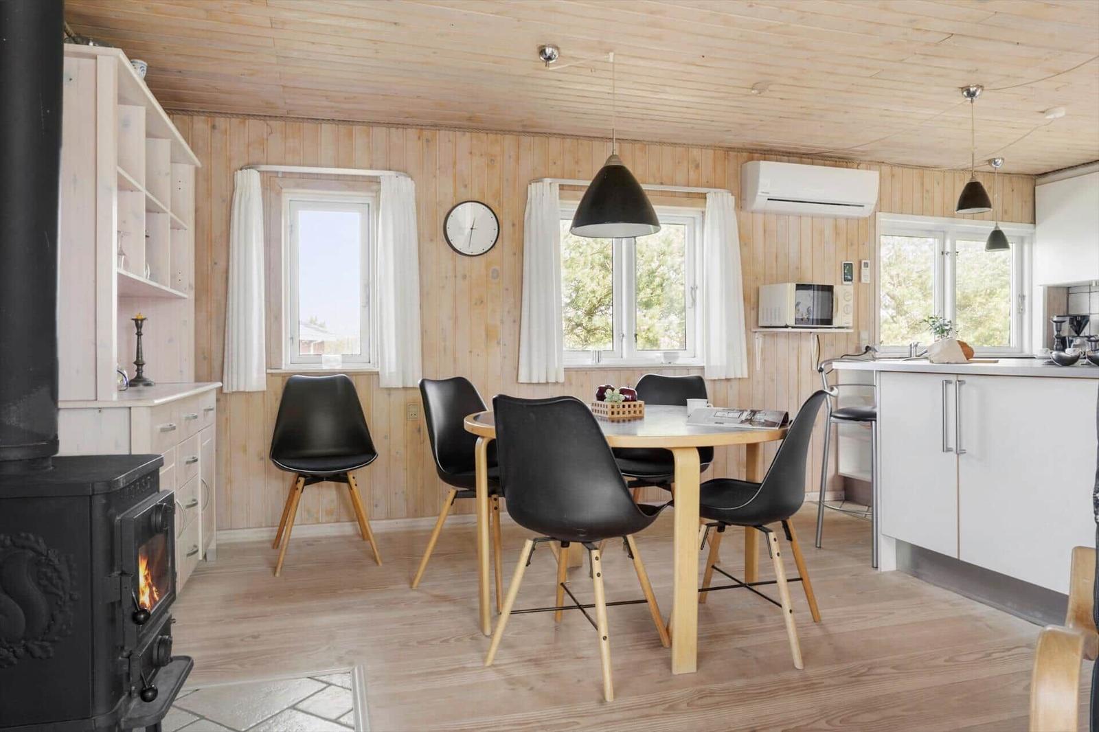 Essbereich mit Tisch, Stühlen und Küchenzeile. Holzwände, Fenster und Kaminofen.