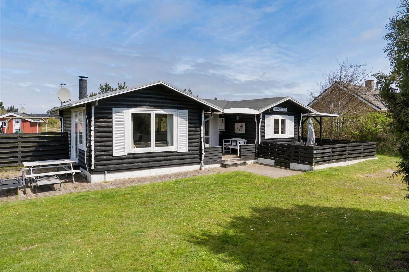 Schwarze Holzhütte mit weißem Fensterrahmen und Terrasse. Grasfläche und Picknicktisch vorne.