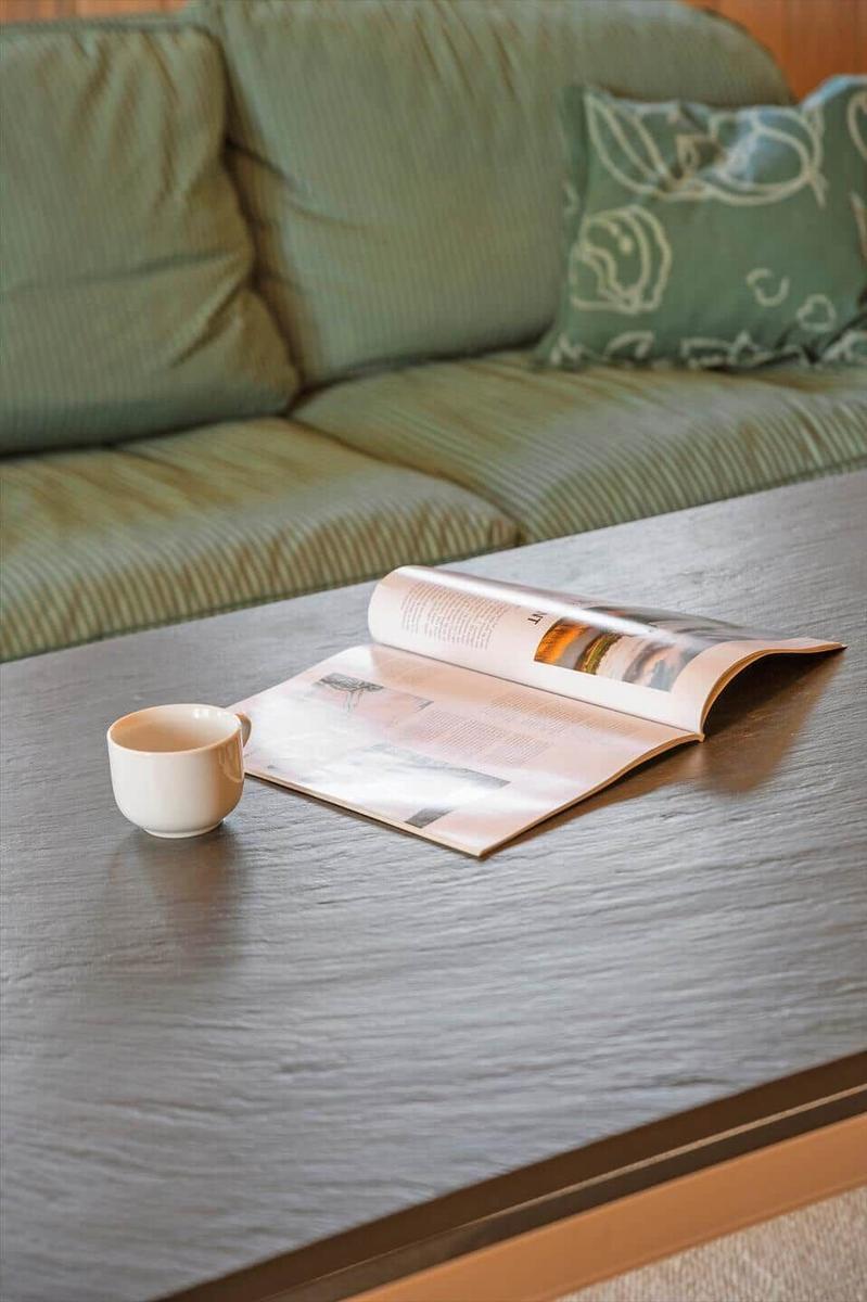 Auf dem Tisch liegt eine offene Zeitschrift und eine Tasse. Im Hintergrund ist ein Sofa mit Kissen.