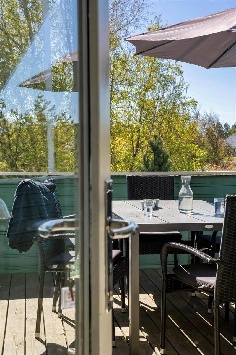 Terrasse mit Tisch, Stühlen und Sonnenschirm. Blick auf grüne Bäume. Holzdecke und Glasfront.