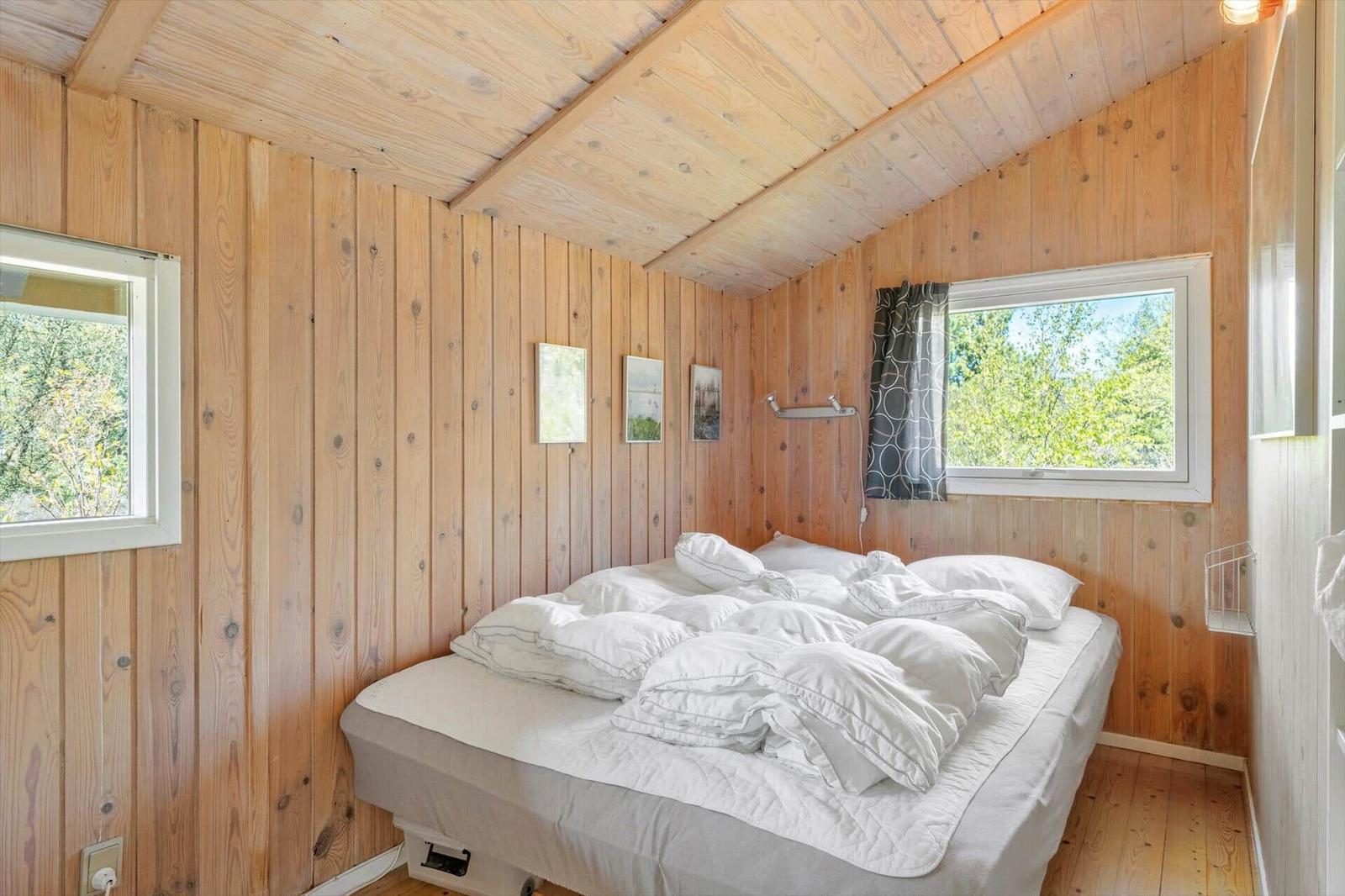 Schlafzimmer mit Holzwänden, Bett und Fenstern mit Blick auf Bäume.