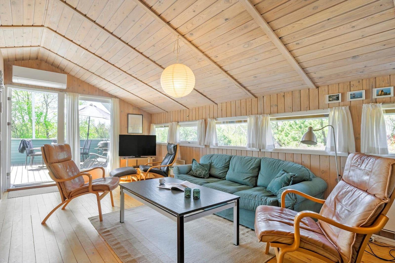 Wohnzimmer mit Sofa, zwei Sessel und Tisch. Holzdecke und Fenster mit Blick auf Außenbereich.