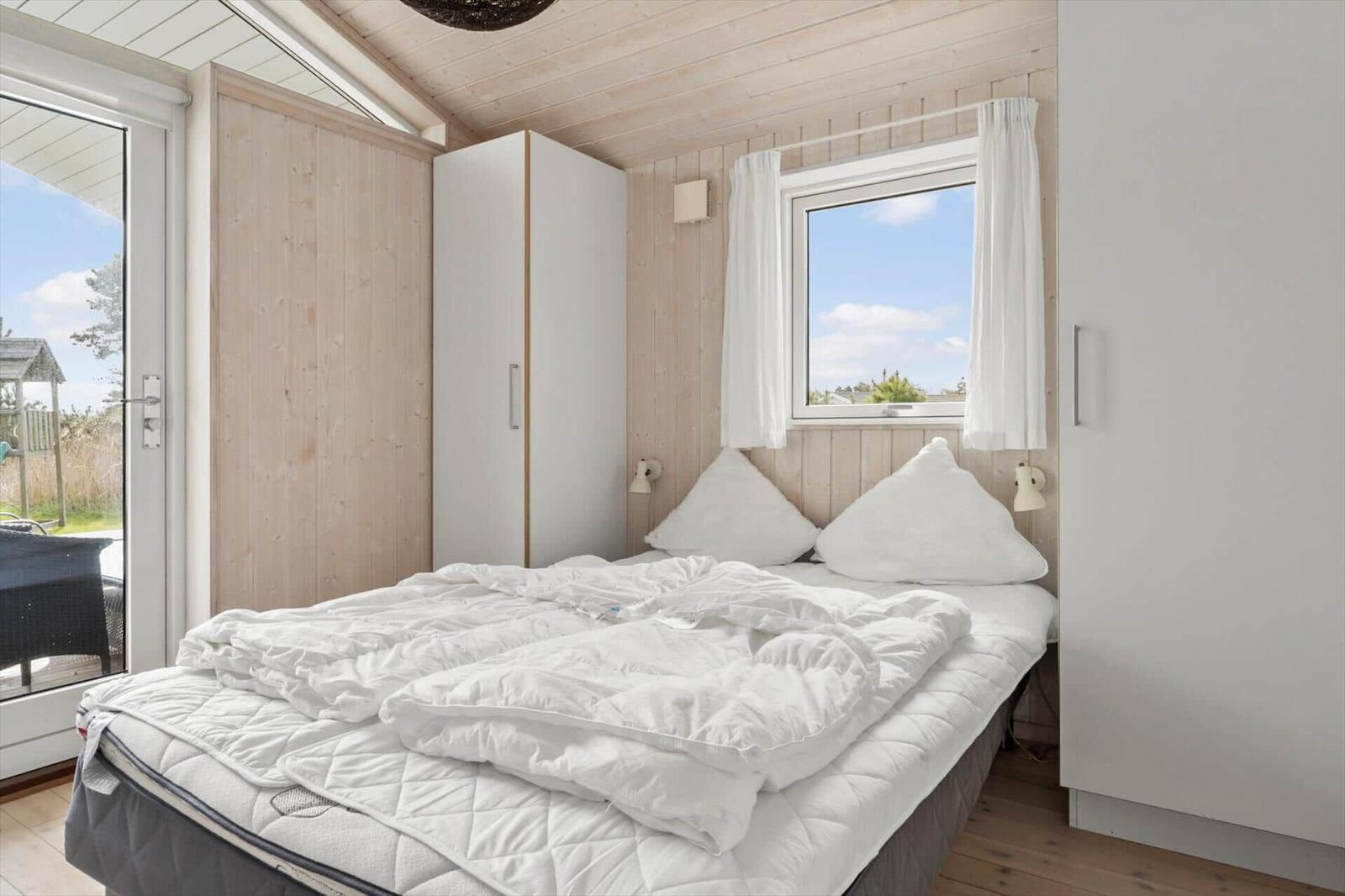 Schlafzimmer mit weißem Bett, Holzwand und Fenster. Tür führt auf Balkon.