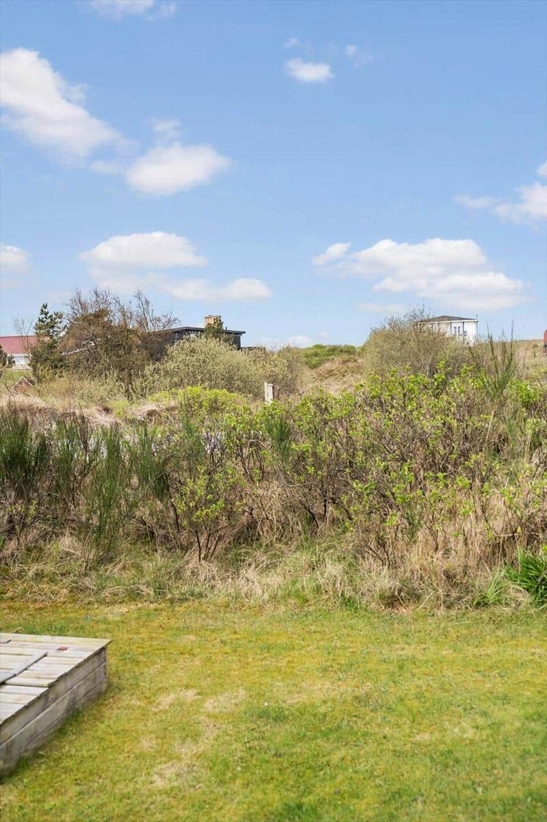 Grüner Rasenbereich mit Holzterrasse und Blick auf Dünenvegetation und Häuser im Hintergrund.