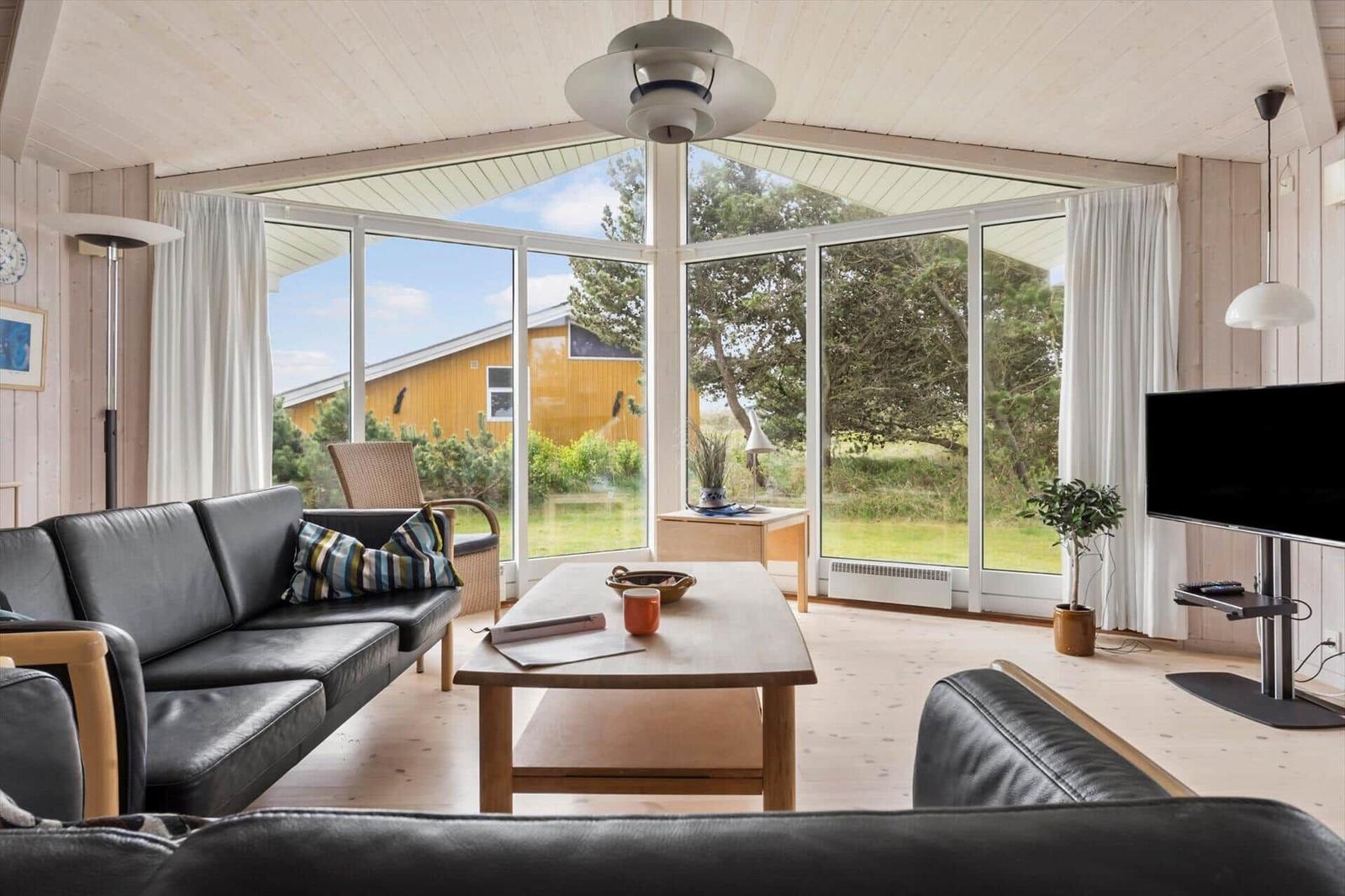 Wohnzimmer mit großem Fenster, Sofa und Couchtisch. Draußen Grün und ein gelbes Haus.