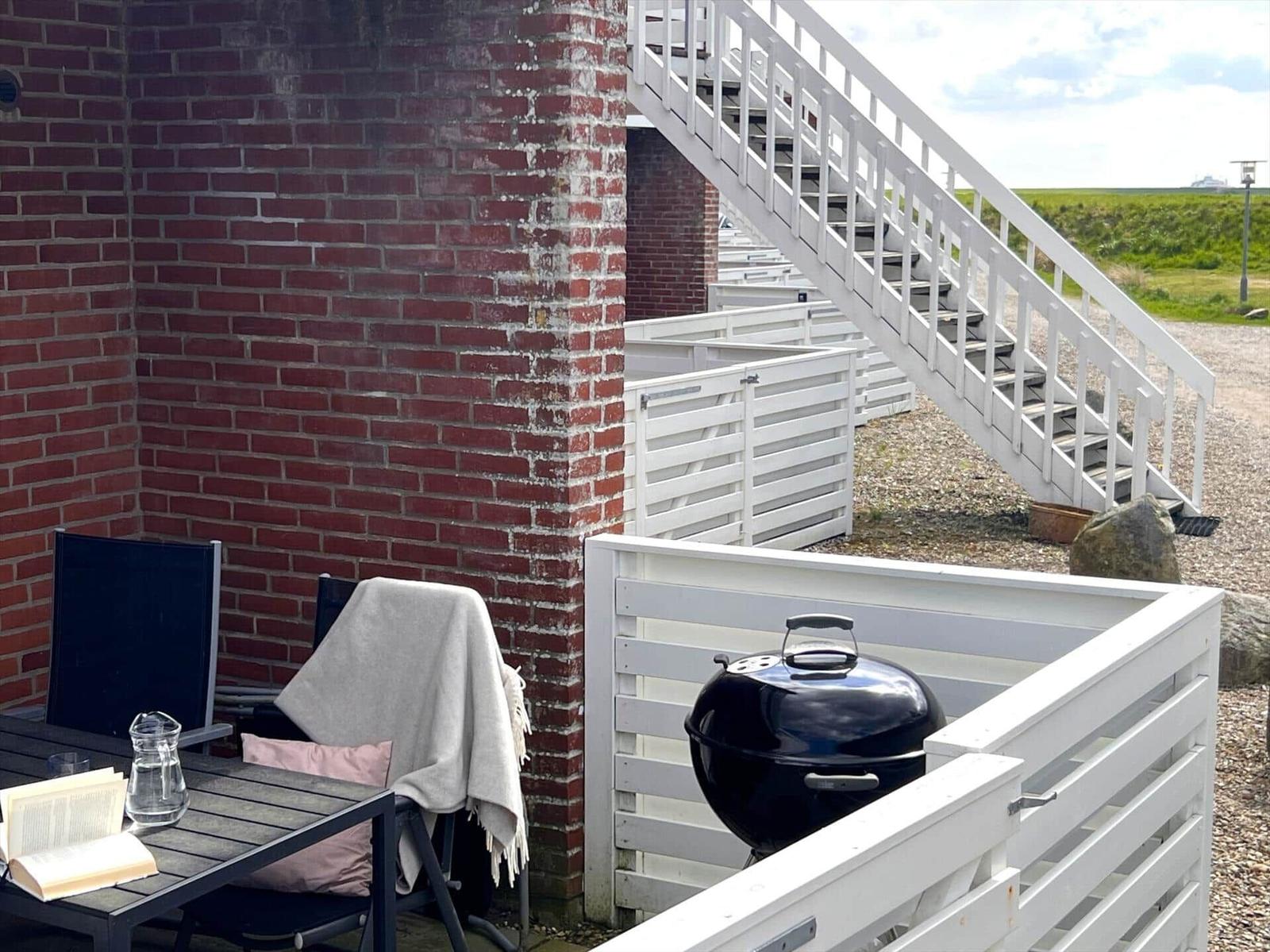 Außenbereich mit Tisch, Stühlen, Grill und rotem Backsteinmauerwerk. Weiße Treppe führt nach oben.