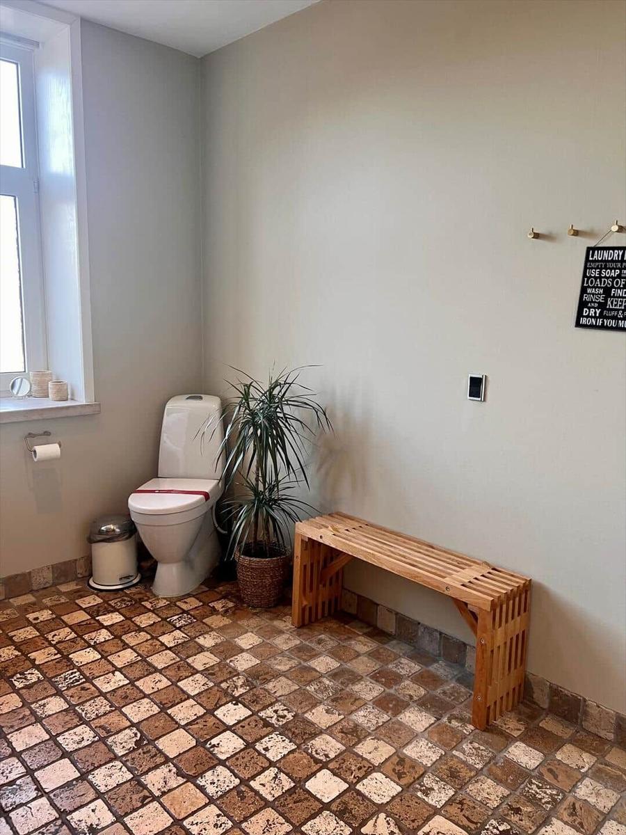 Toilette mit Holzbank, Pflanze und Fenster. Mosaikboden und Wandhalter.