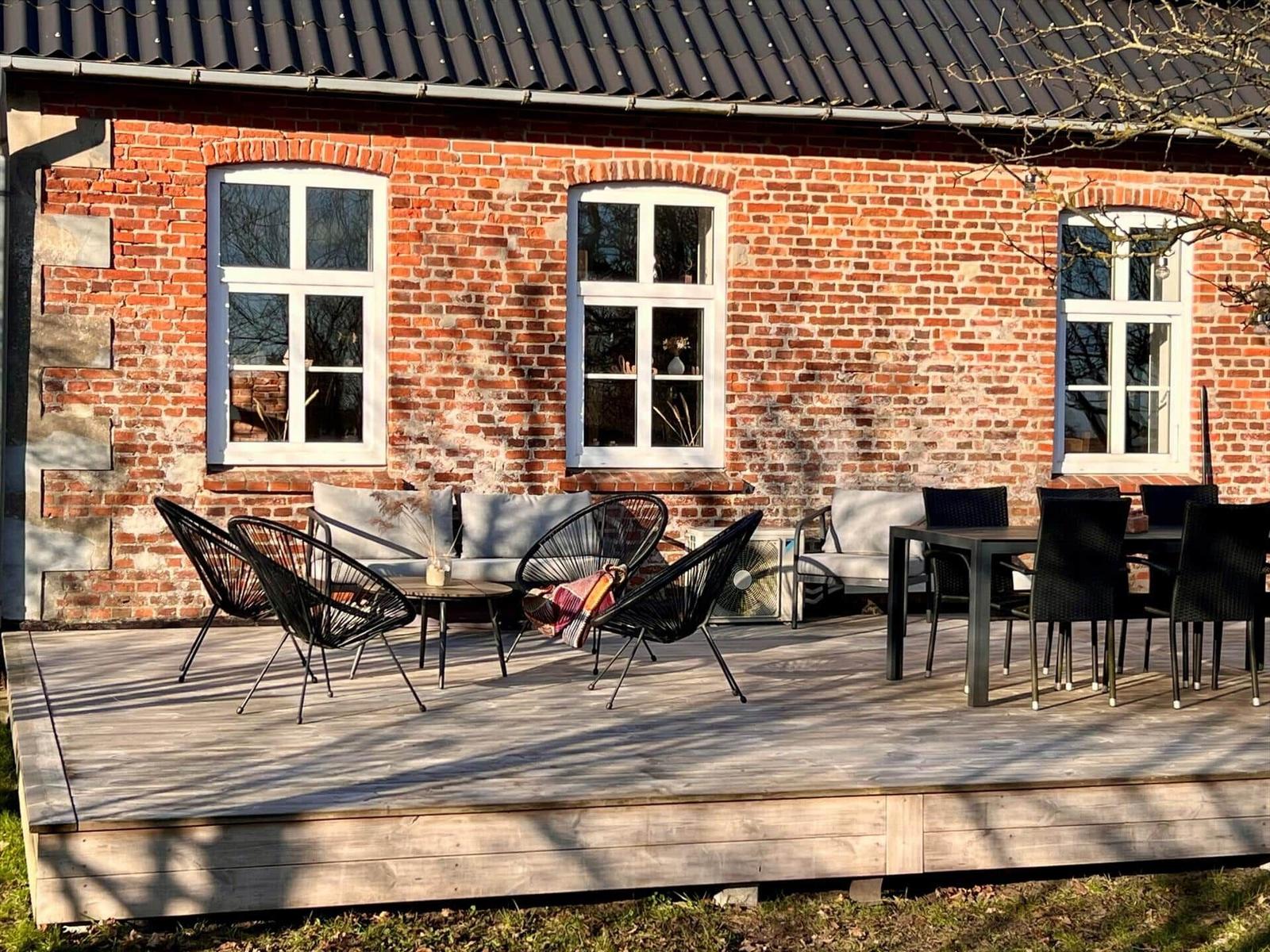Holzterrasse mit Sitzgruppe und Tisch vor Backsteinhaus mit weißen Fenstern.
