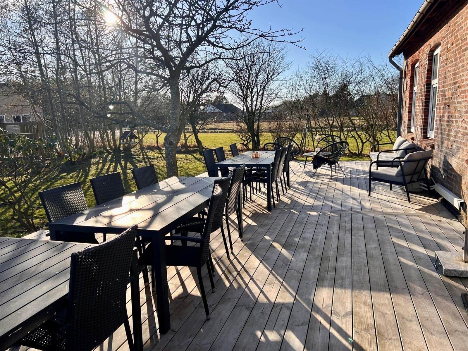Holzterrasse mit Tischen und Stühlen, Blick auf Grünfläche und Backsteinhaus.