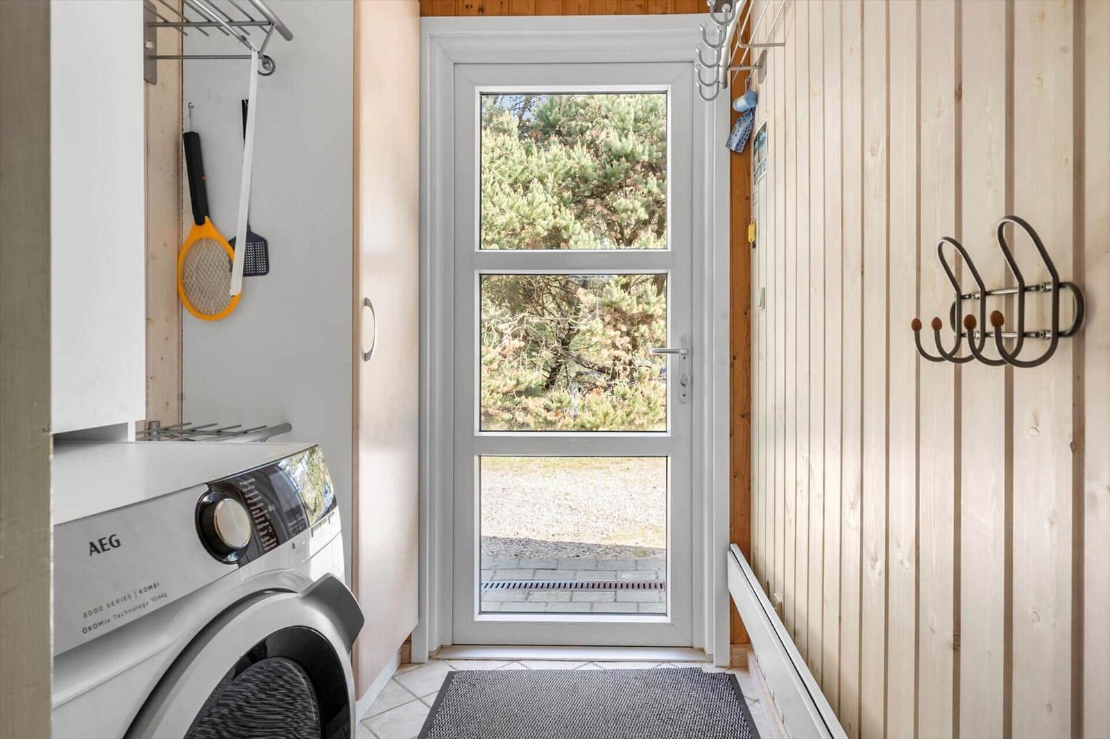 Eingangsbereich mit Waschmaschine und Holzwand. Durch das Fenster ist ein Wald sichtbar.
