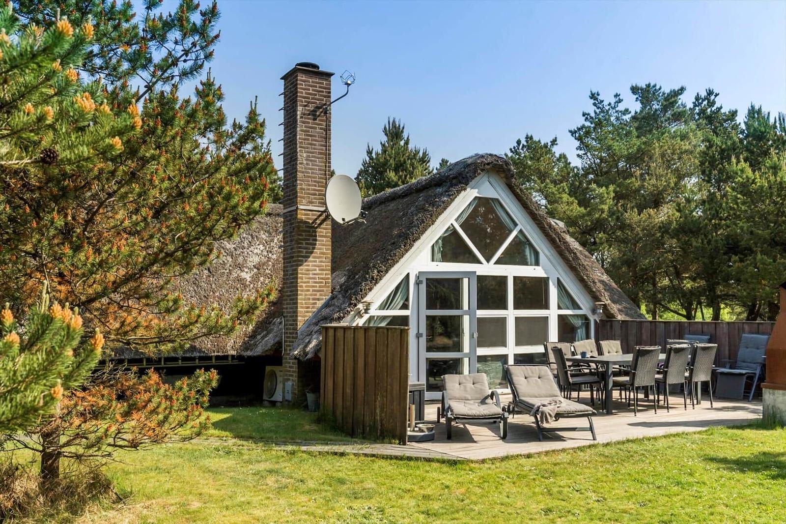 Haus mit Strohdach, Terrasse mit Liegen und Tisch, umgeben von Nadelbäumen.