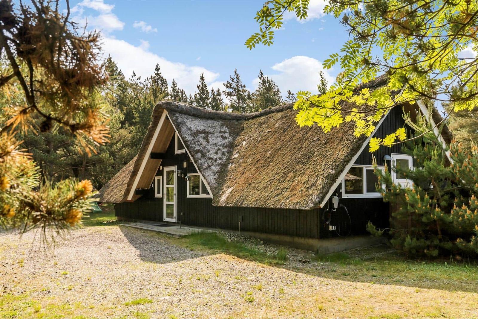 Ein schwarz gestrichenes Haus mit Strohdach steht im Wald. Vor dem Haus befindet sich ein Kiesweg.