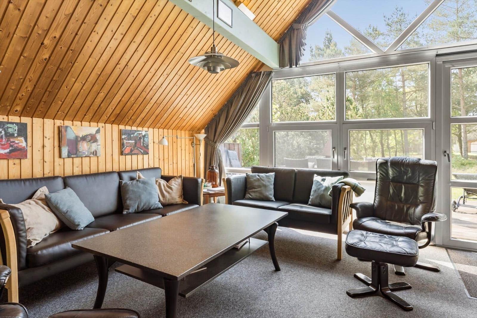 Wohnzimmer mit Holzdecke, Sofa, Sessel und großem Fensterblick auf Wald.