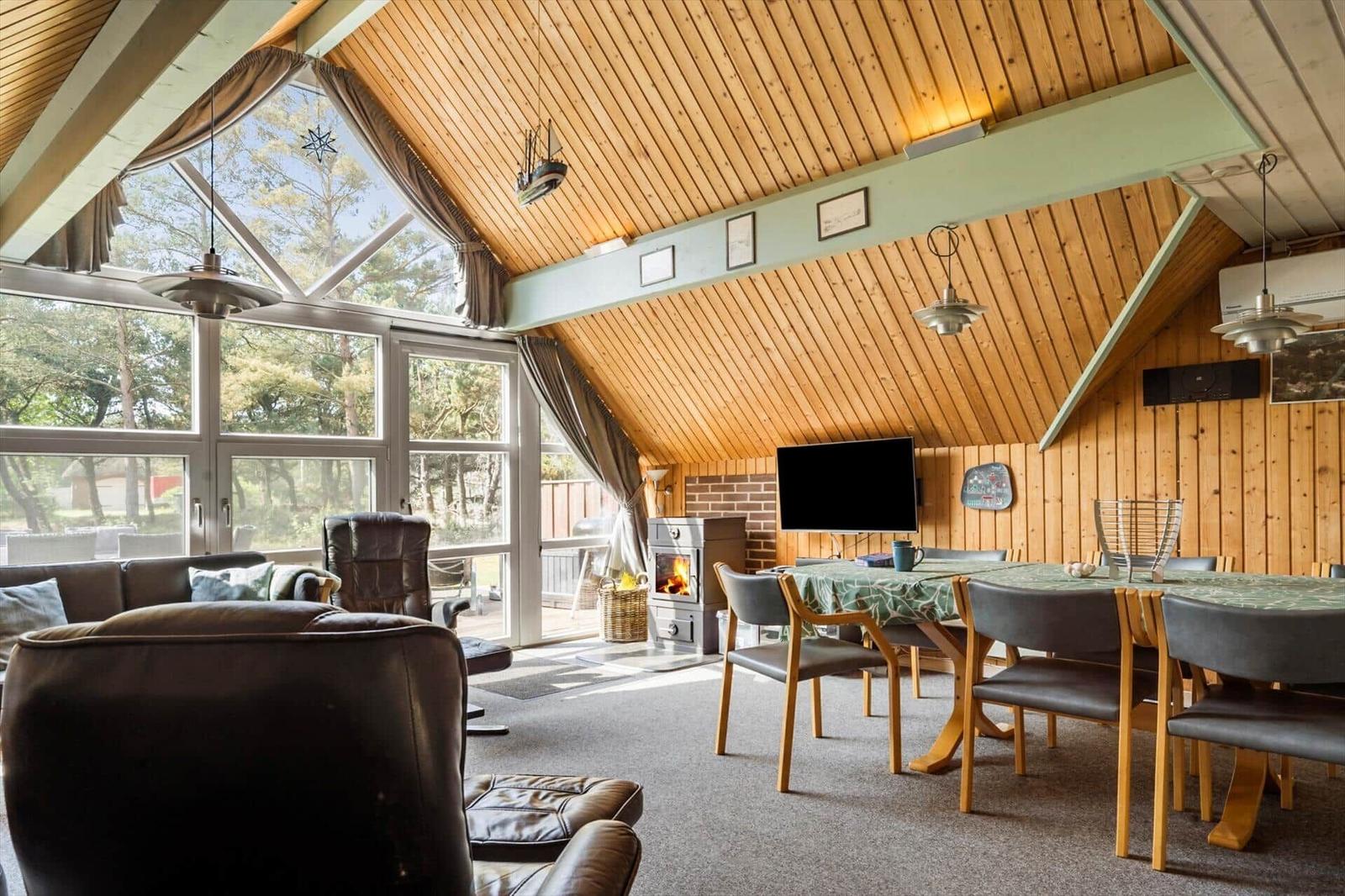 Wohn- und Essbereich mit Holzdecke, Kamin und großem Fensterblick auf den Garten.