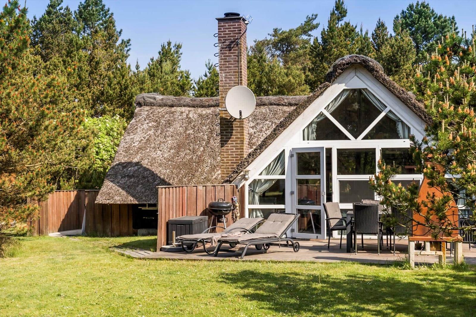 Haus mit Strohdach, Terrasse mit Liegen und Grill, umgeben von Bäumen.