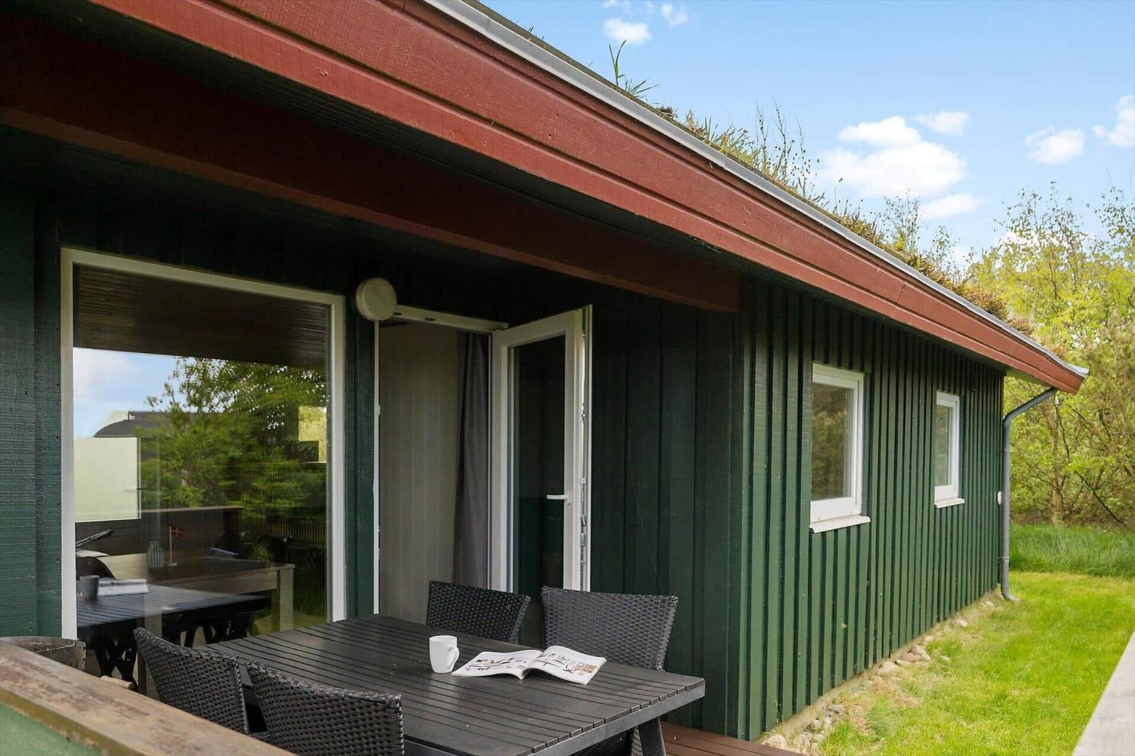 Grünes Holzhaus mit Terrasse, Tisch, Stühlen und Grasdach unter blauem Himmel.