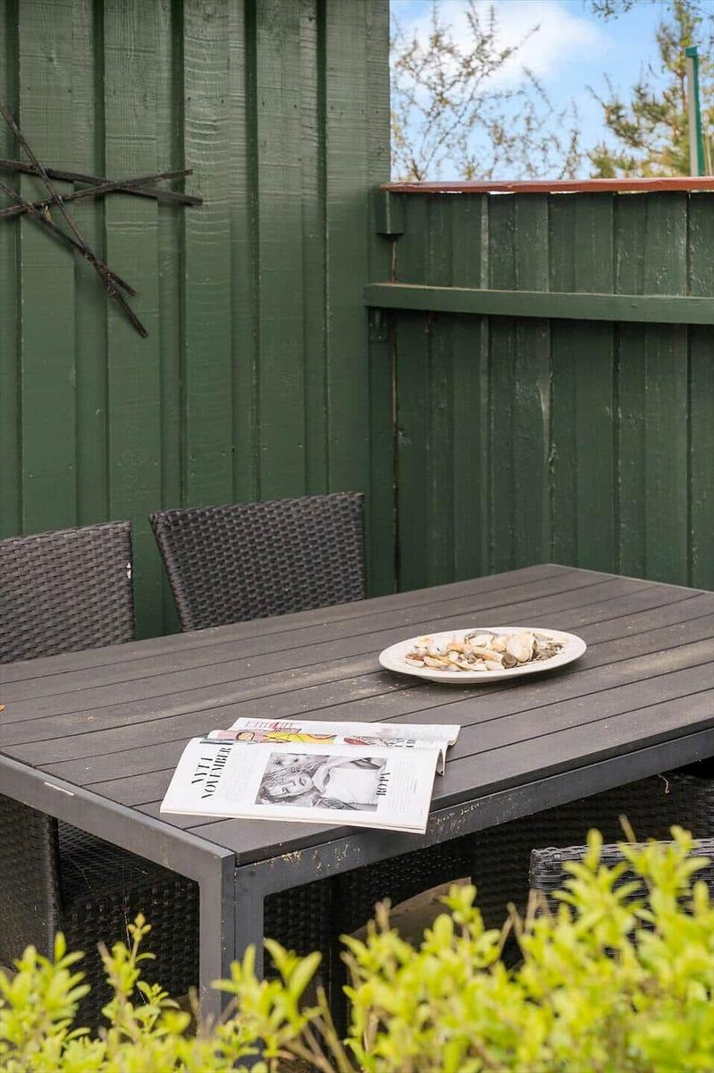 Holztisch mit Stühlen, Teller und Zeitung im Garten. Grüne Holzwand im Hintergrund.