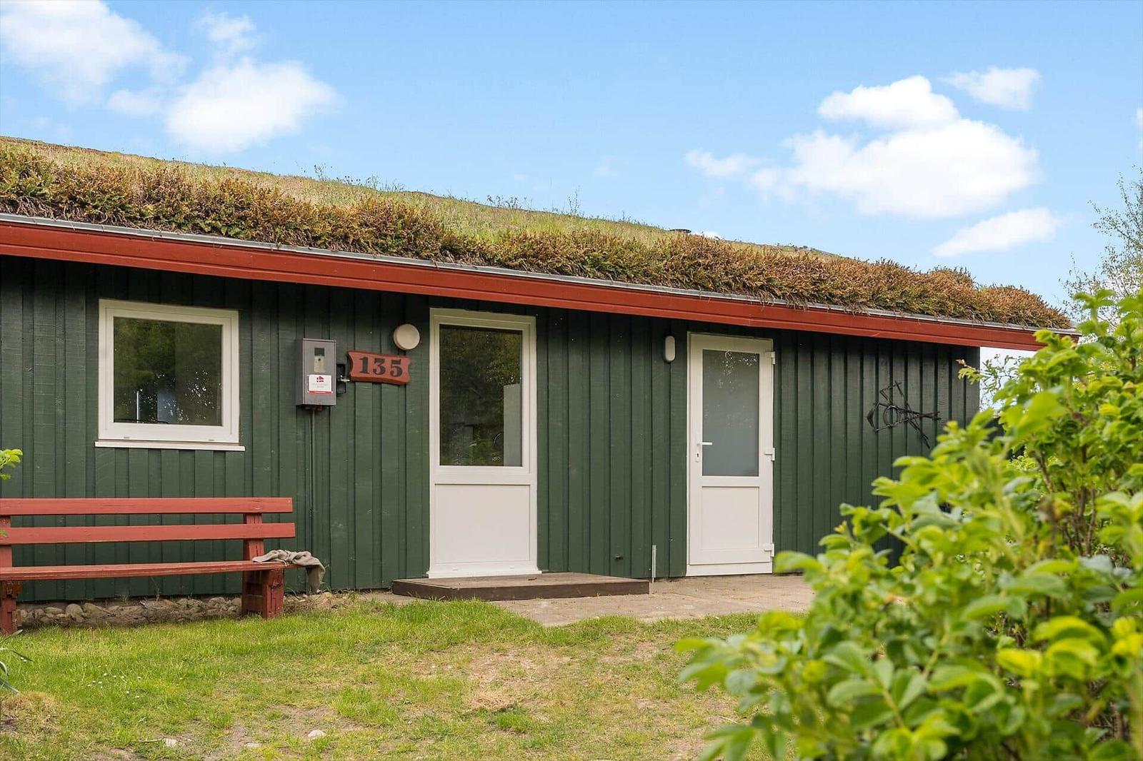Ein grünes Holzhaus mit grasbedecktem Dach, zwei weißen Türen und einer roten Bank.