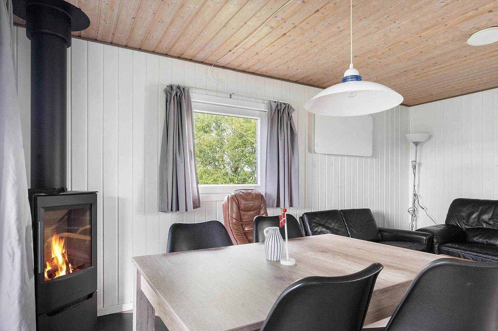 Essbereich mit Tisch, Stühlen und Kamin. Fenster mit grauen Vorhängen. Holzdecke und weiße Wände.