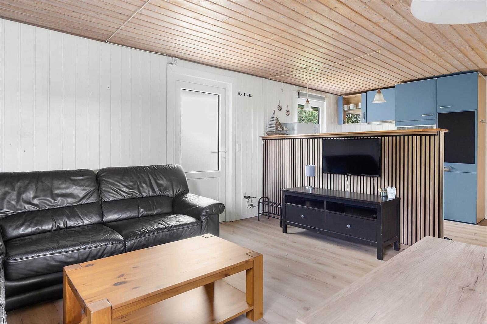 Wohnzimmer mit Ledersofa, Holztisch und TV. Küchenbereich mit blauen Schränken.