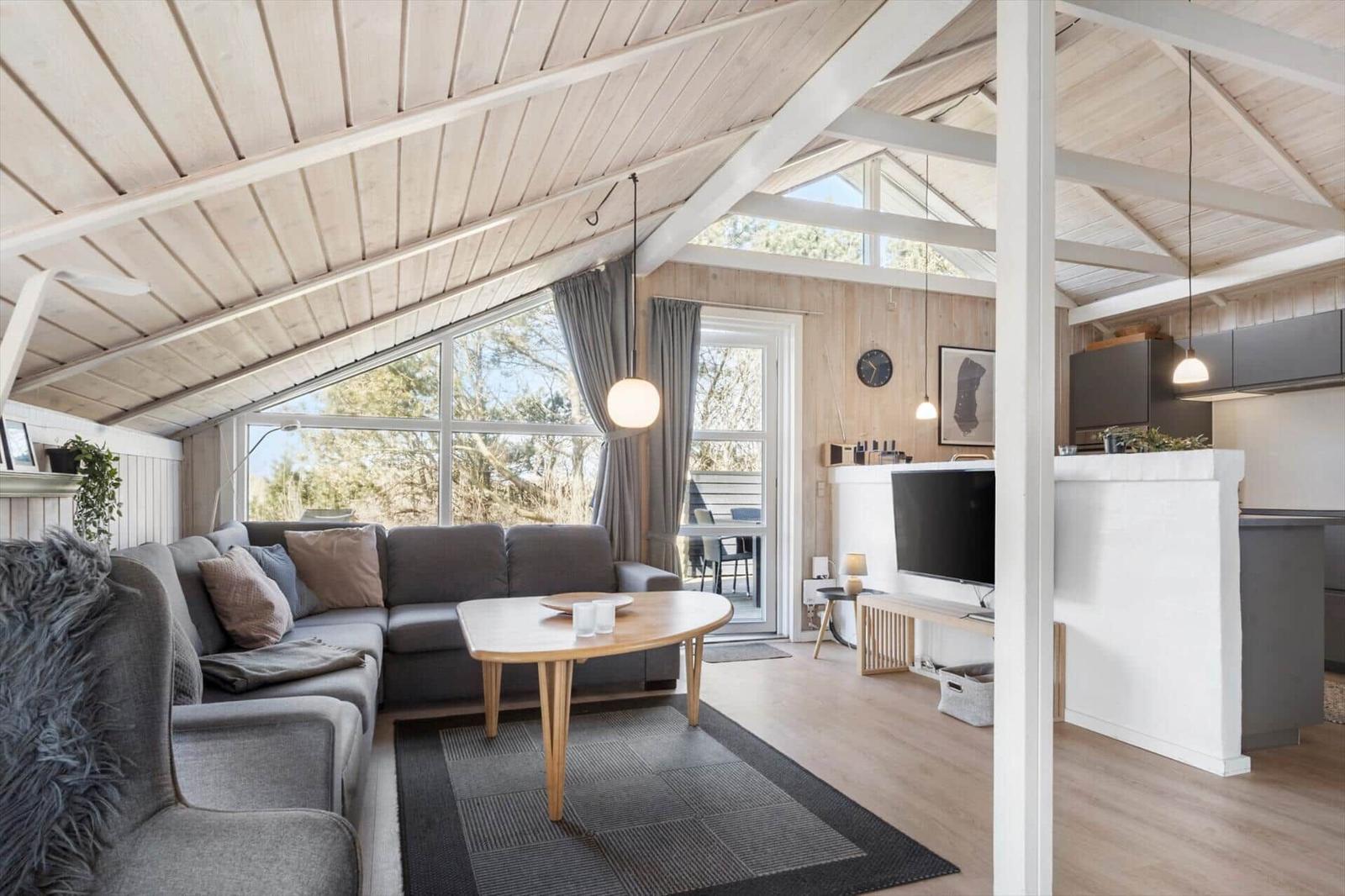 Wohnbereich mit Sofa, Tisch und TV. Küche mit Arbeitsplatte. Holzdecke und große Fenster.