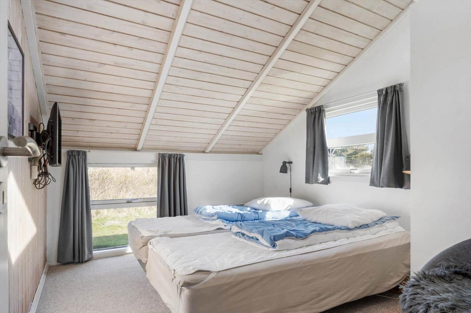 Schlafzimmer mit Doppelbett, Holzdecke und Fenstern mit grauen Vorhängen.