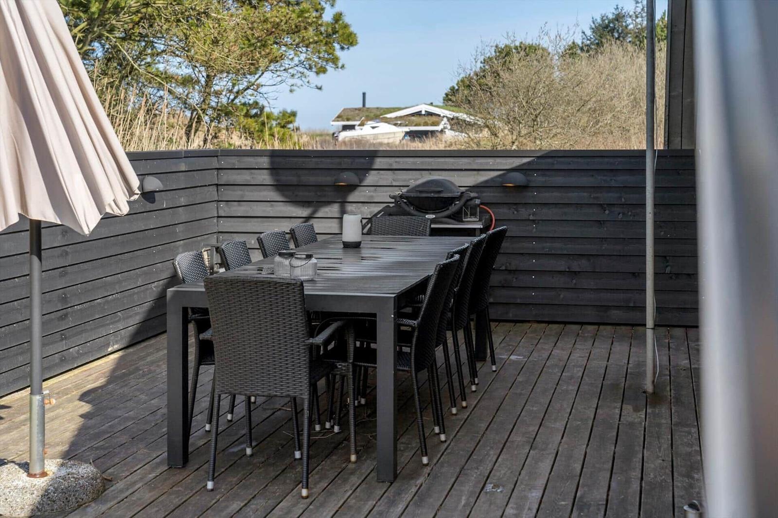 Terrasse mit Tisch, Stühlen und Grill. Hintergrund: Holzwand und Bäume.