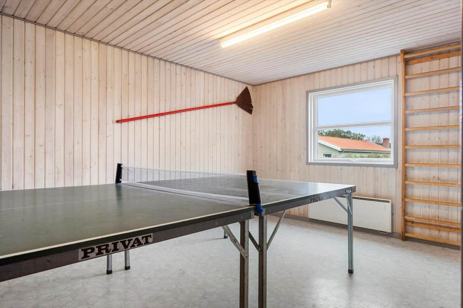 Tischtennisplatte in einem Raum mit Holzwänden und Fenster. An der Wand hängt eine Besenstange.
