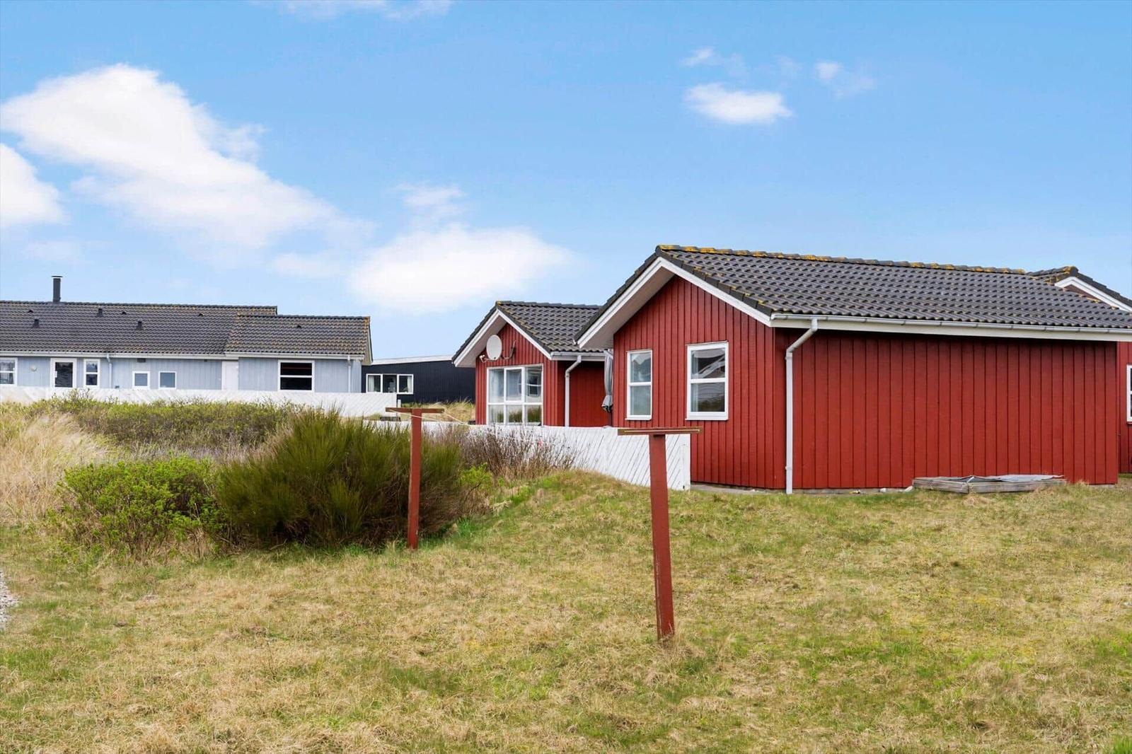Rotes Ferienhaus mit Dach und Fenstern, umgeben von Gras und Sträuchern unter blauem Himmel.