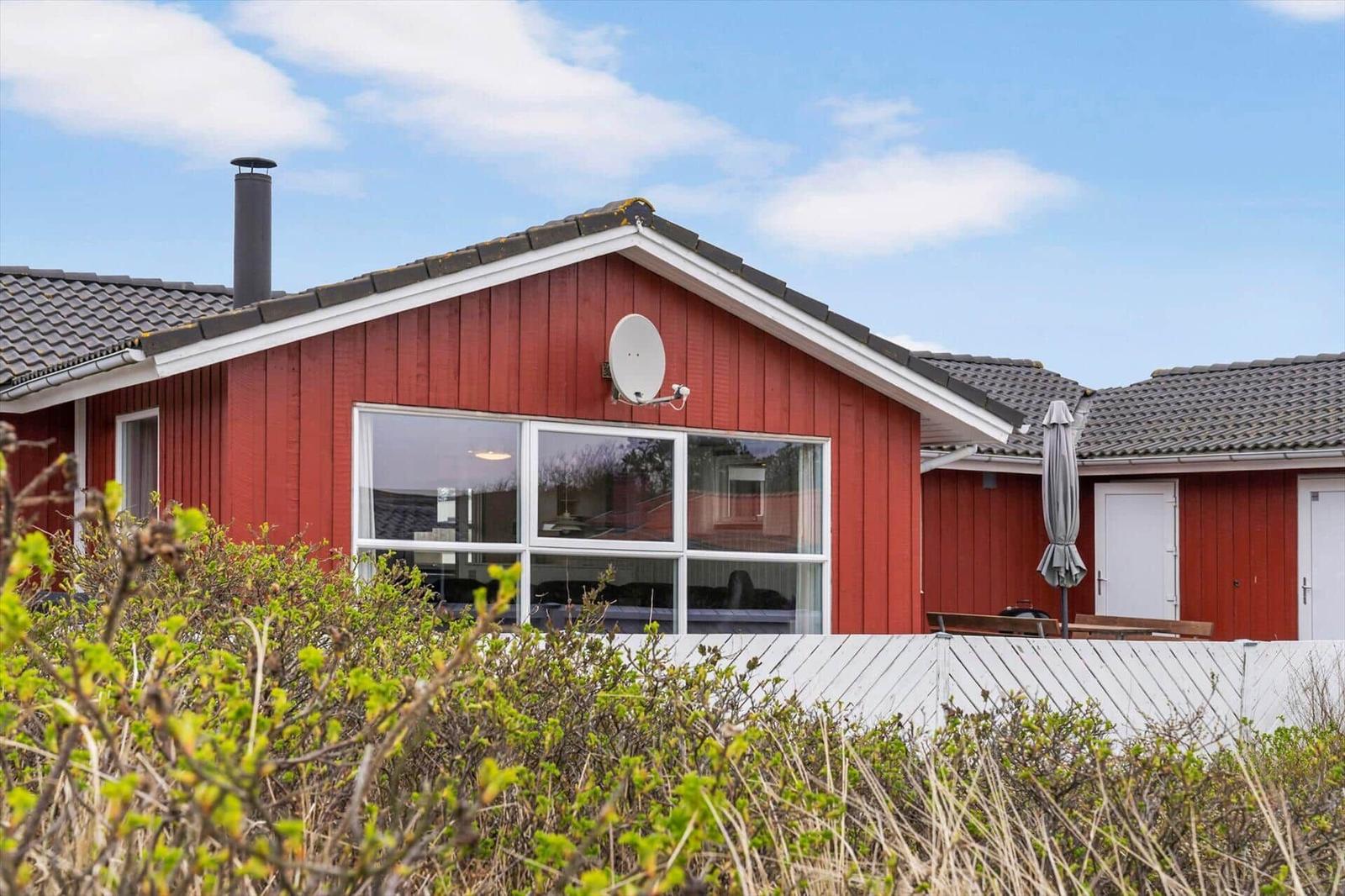 Rotes Ferienhaus mit großem Fenster und Satellitenanschluss. Außenbereich mit Sonnenschirm und Tisch.
