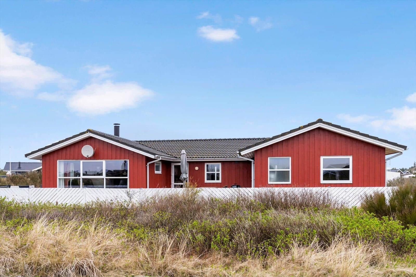 Rotes Ferienhaus mit weißem Zaun und Garten. Satellitenschüssel und Fenster sichtbar.
