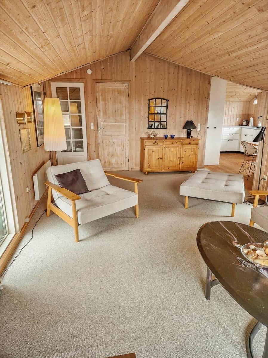 Wohnzimmer mit Holzwand, Sessel, Couchtisch und Küchenbereich im Hintergrund.