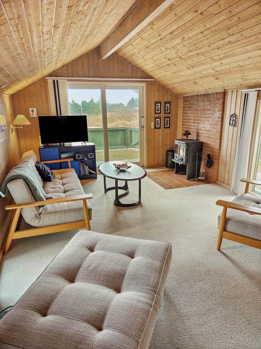 Wohnzimmer mit Holzdecke, Sofa, Sessel, Kamin und TV. Blick auf Terrasse.