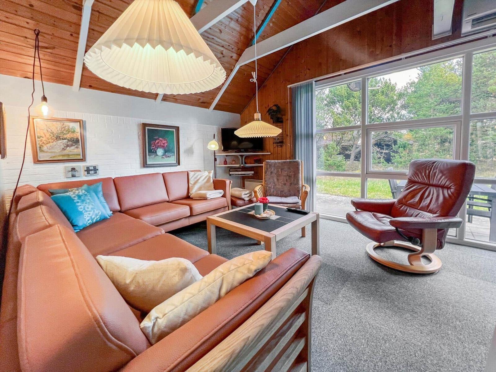 Living room with brown sofa, armchair, and large window. Wooden ceiling and wall pictures.