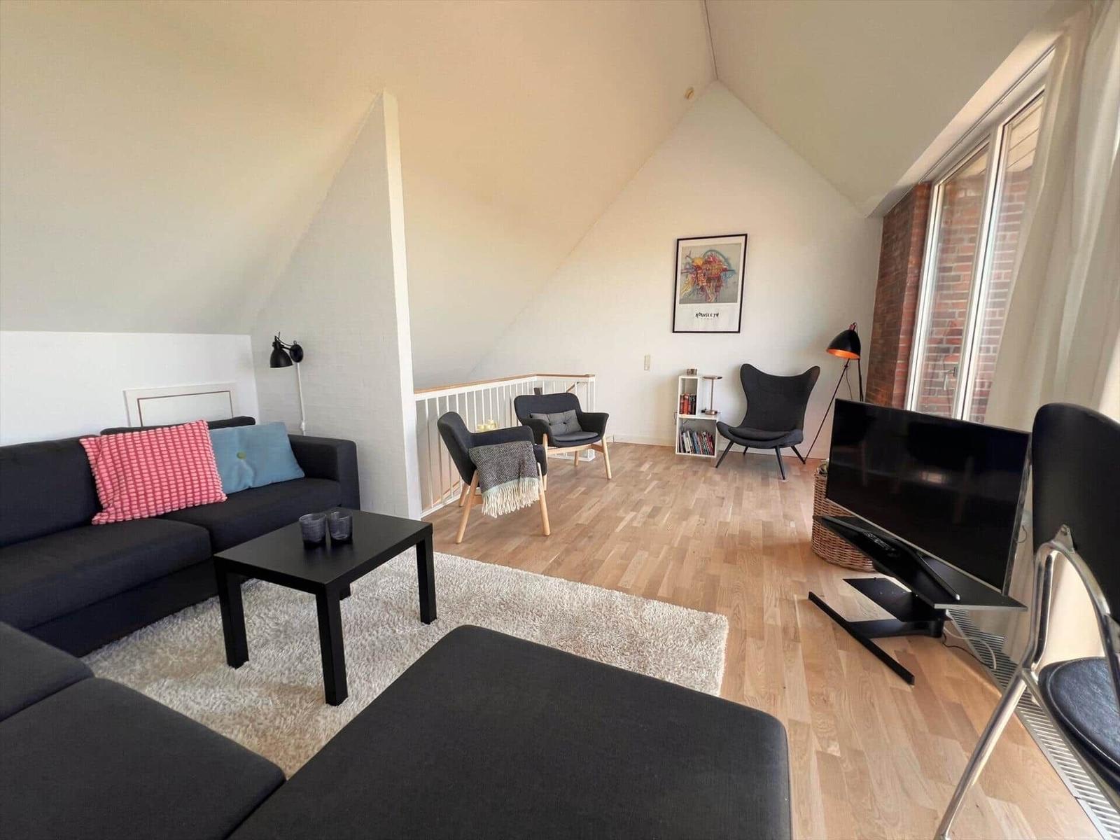 Living room with sofa, chairs, and TV. Wooden floor and large windows.