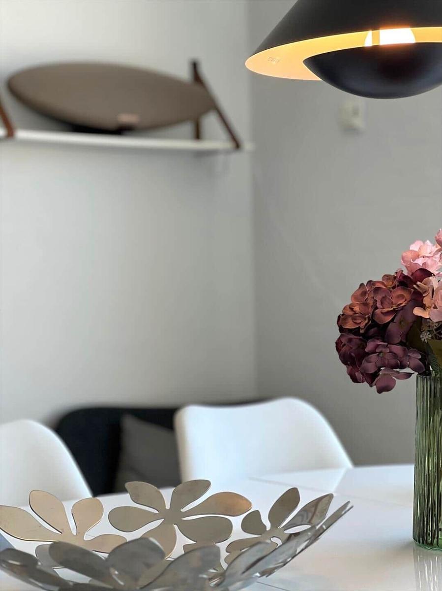 Dining table with flower vase and modern lamp. Background: chair and wall shelf.