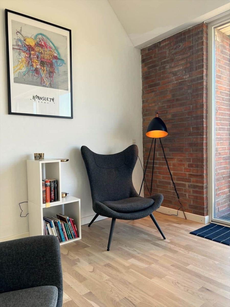 A dark chair stands before a brick wall. Beside it is a floor lamp and a bookcase.